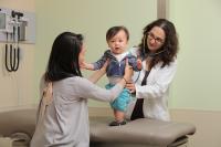Female physician examines toddler while mother holds him in standing position.